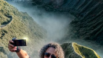 A long-haired man takes a selfie with a hilltop background
